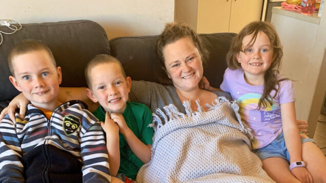 Terri Grosser at home on the sofa with her children. Terri and her children are smiling for the camera. Terri has a blanket on top of her.