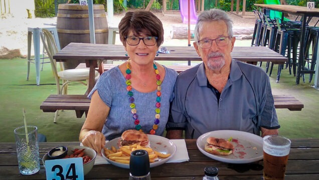 Jackie and her husband, Bob, sit at a picnic table eating food. Jackie is wearing a wig after cancer treatment