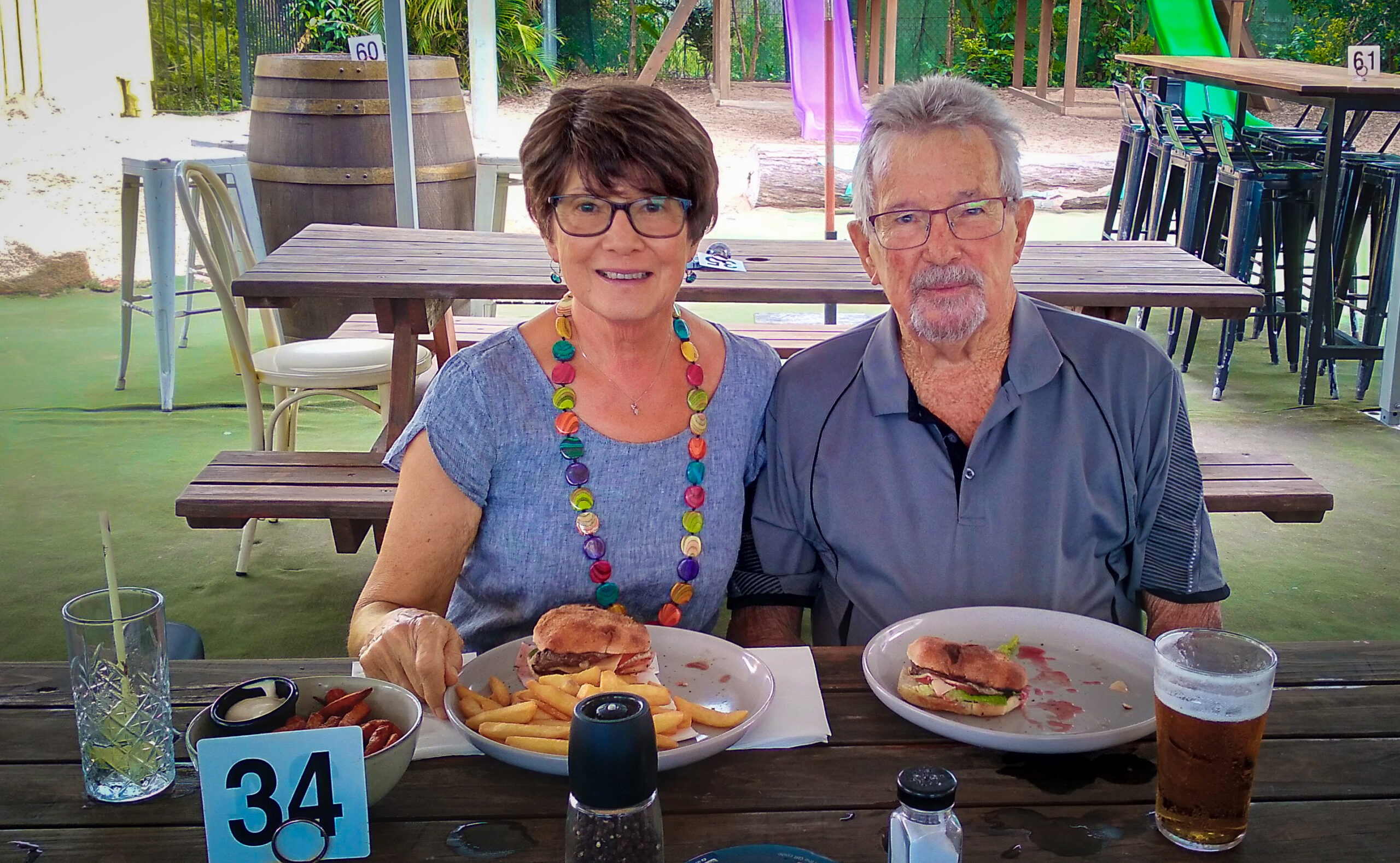 Jackie and her husband, Bob, sit at a picnic table eating food. Jackie is wearing a wig after cancer treatment