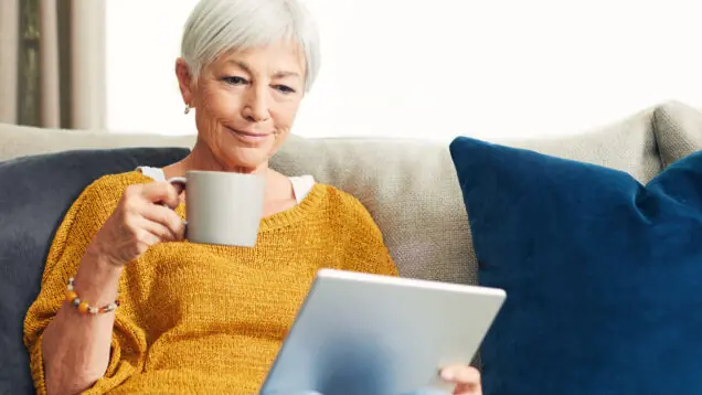 A white woman with white hair sits on a sofa with a mug of tea reading about palliative care on a digital tablet