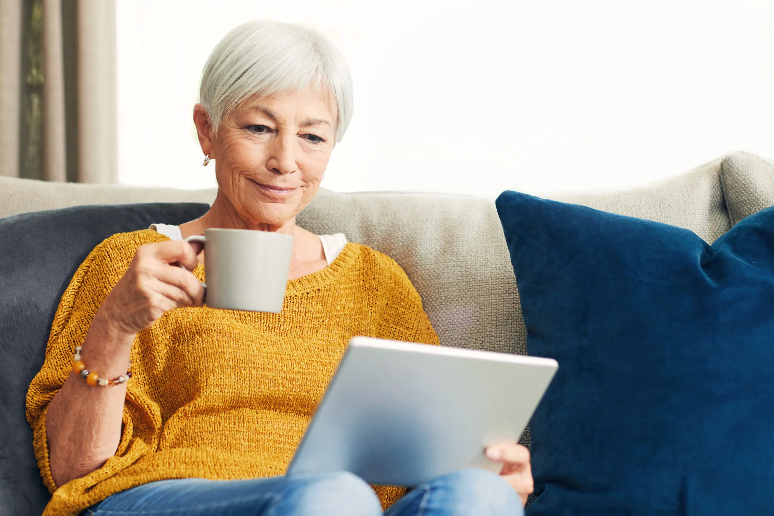 A white woman with white hair sits on a sofa with a mug of tea reading about palliative care on a digital tablet